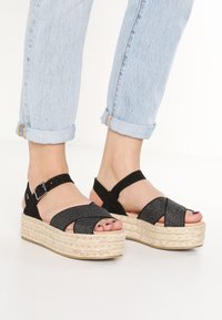 Czarne sandały espadryle z teksturowaną cholewką, plecioną platformą z juty oraz regulowanym paskiem na kostkę; zaprojektowane w stylu z krzyżującymi się paskami.