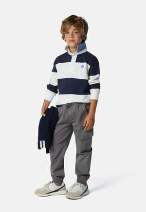 Marineblaues und weiß gestreiftes Rugbyshirt mit Kragen und Knopfleiste, kombiniert mit grauen Cargo-Hosen und cremefarbenen Sneakers. Hält einen marineblauen Pullover.
