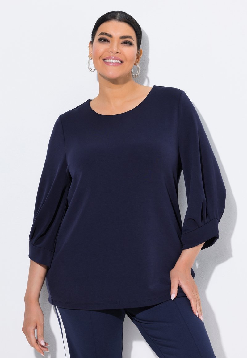 Laivastonsininen paita, jossa on kolmen neljänneksen puffihihat ja pyöreä pääntie, valmistettu sileästä kankaasta, joka hohtaa hienovaraisesti. Peplum-tyylinen helma.