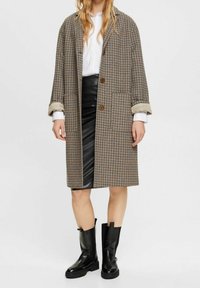 Cappotto in motivo houndstooth marrone con grandi revers, due bottoni e maniche arrotolate, abbinato a una gonna in pelle nera e stivali neri chunky.