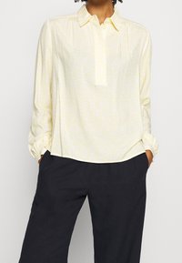 Lichtgele blouse met subtiel bloemenpatroon, met kraag, knoopsluiting en lange mouwen, gecombineerd met donkere broek.
