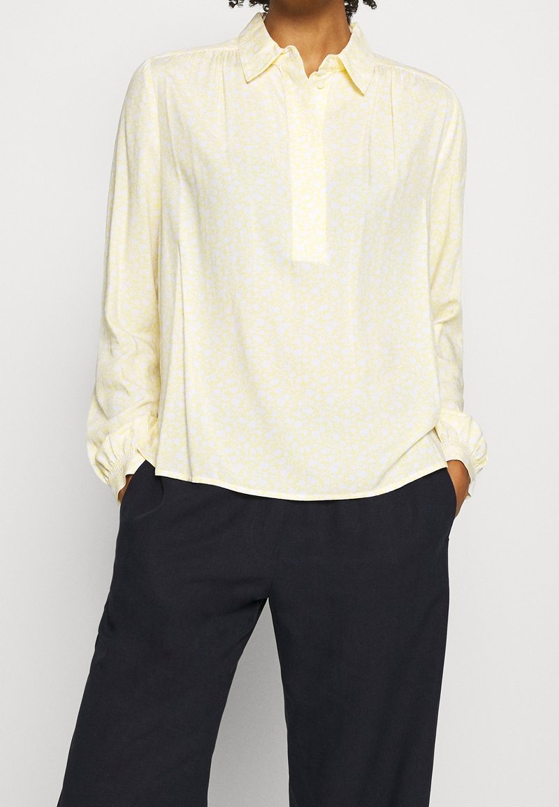 Lichtgele blouse met subtiel bloemenpatroon, met kraag, knoopsluiting en lange mouwen, gecombineerd met donkere broek.