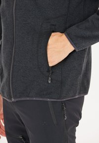 Giacca in pile grigio scuro con tasche con zip, caratterizzata da una texture liscia e cerniere nere a contrasto. Design aderente con branding minimale.
