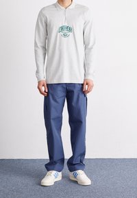 Grå långärmad polo med dragkedja och grön logotyp, kombinerad med blå cargobyxor och vita sneakers med blå ränder.