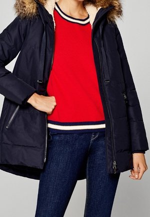 Osoba ubrana w granatową parkę z kapturem wyłożonym futrem, na swetrze czerwonym z białymi i granatowymi paskami, do tego ciemnoniebieskie dżinsy.