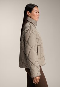 Giacca trapuntata beige con colletto alto, tasche laterali e silhouette ampia. Il materiale sembra morbido con una finitura strutturata.