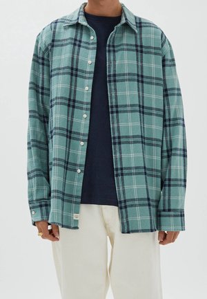 Man draagt een teal en marineblauw geruit overhemd over een zwart T-shirt, gecombineerd met off-white broek, staand tegen een effen achtergrond.