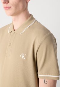 Bežs polo krekls ar teksturētu audumu. Tam ir balta apdare apkaklīte un mazs balts logo kreisajā krūtī. Klasiska divu pogu aizdare.