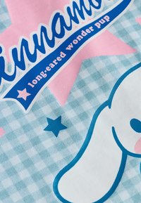Ljusblå rutig tyg med en stor stiliserad vit hund, rosa detaljer, och blå text som säger "Cinnamoroll långörat underverkshund."