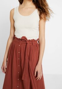 Débardeur blanc côtelé associé à une jupe longue couleur rouille avec une taille à volants et des boutons sur le devant.