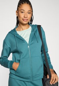 Τζελ τζάκετ με φερμουάρ, κουκούλα και μπροστινές τσέπες, φτιαγμένο από απαλό υλικό, συνδυασμένο με πουκάμισο ανοιχτού πράσινου χρώματος, με άνετη εφαρμογή.