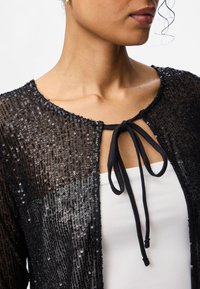 Svart paljettbeströdd cardigan med tajt passform, utrustad med rund halsringning och knytning framtill i svart tyg. Texturerad yta.