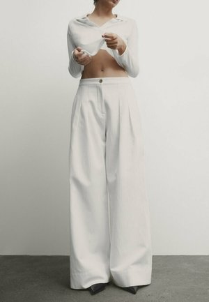 Witte cropped top met lange mouwen en een kraag, gecombineerd met witte, wijde, hoog getailleerde broeken met een knoopsluiting aan de voorkant en plooien.