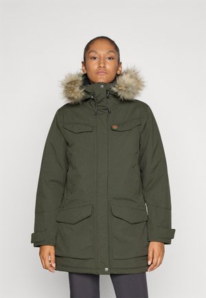 Vrouw in een olijfgroene parka met een gevoerde capuchon van bont, voorzakken met klep en ritsopening, staand tegen een effen lichte achtergrond.