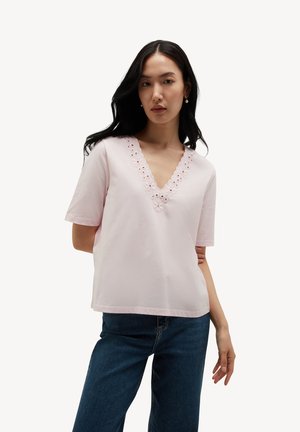 Mujer con cabello largo y oscuro que lleva una camiseta de manga corta de color rosa claro con escote en V bordado y vaqueros azules sobre un fondo blanco liso.