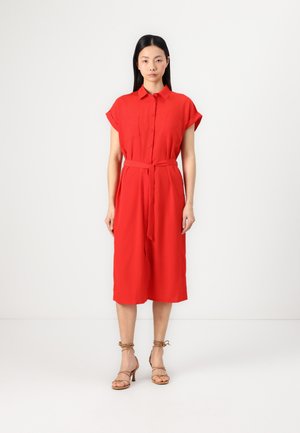Robe rouge à boutons avec manches courtes, deux poches poitrine et ceinture à la taille. Texture lisse, longueur au genou, accompagnée de talons à bride de cheville beige.