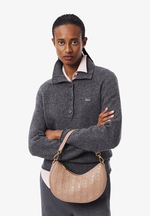 Maglione grigio lavorato a maglia con collo alto, chiusura a bottoni e una camicia rosa chiaro sotto; tiene in mano una borsa in pelle texturizzata color rosa antico.