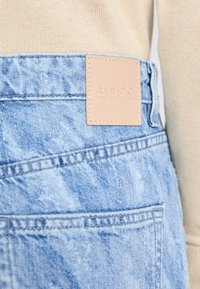 Calções de ganga em azul desbotado com um acabamento texturizado, apresentando um rótulo de couro bege com "BOSS" estampado e detalhes de costura em contraste.