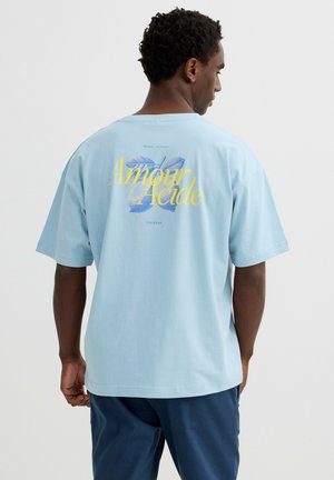 Hombre con camiseta azul claro con texto amarillo "Amour Acide" y gráfico de hoja azul en la espalda, combinado con pantalones azul oscuro.