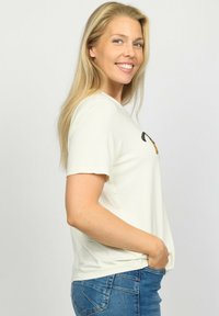 Lys cremefarvet kortærmet t-shirt med rund hals, med et dekorativt design i sort og guld på forsiden. Sammen med blå jeans.