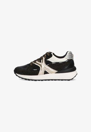 Zwart-witte sneaker met een glanzend gouden accent, een gestructureerde mesh bovenkant, een dikke witte zool en een zwarte rubberen loopzool.