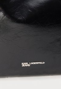 Czarna fakturowana powierzchnia ze skóry z srebrnym logo marki "KARL LAGERFELD JEANS" umieszczonym centralnie blisko dolnej krawędzi.