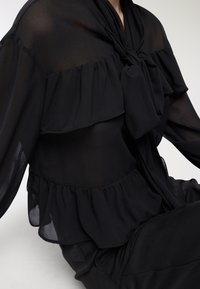 Blusa nera trasparente con un design arricciato sul petto e in vita, caratterizzata da un collo annodato e maniche lunghe, realizzata in tessuto leggero.