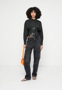 Czarna krótka koszula z błyszczącym panelem, zestawiona z ciemnymi spodniami bojówkami z kieszeniami. Modelka trzyma jaskrawo pomarańczową torbę. Całość dopełniają proste sandały.