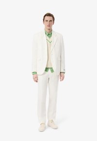 Completo in lino bianco con giacca a un bottone, sovrapposto a una camicia verde e un gilet di maglia crema. Il modello indossa sneakers beige.