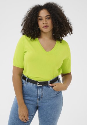 Frau mit lockigem Haar, die ein limettengrünes V-Ausschnitt-Kurzarmshirt, blaue Jeans und einen schwarzen Gürtel vor einem schlichten weißen Hintergrund trägt.