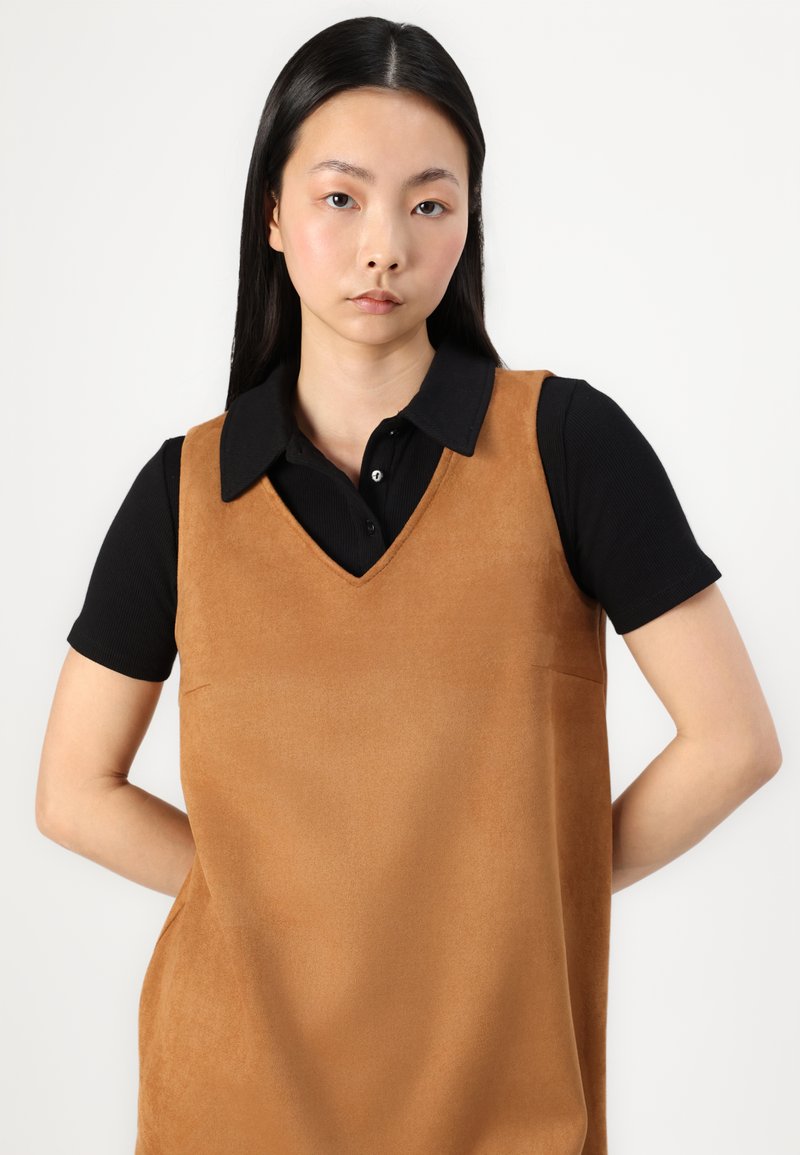 Braunes Wildleder-Sleeveless-Kleid mit V-Ausschnitt, getragen über einem schwarzen Hemd mit Kragen. Glatte Textur, minimalistisches Design und subtile Seitentaschen-Details.