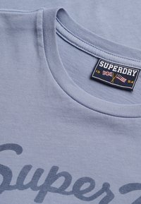 Detalle del cuello de una camiseta azul claro con etiqueta "Superdry" que presenta las banderas del Reino Unido y de Estados Unidos, y texto impreso parcial en la tela.