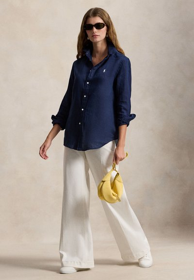 Marineblå lin-skjorte med oppbrettede ermer, hvite vidbukser i denim, og en gul håndveske. Modellen har på seg svarte solbriller og står oppreist.