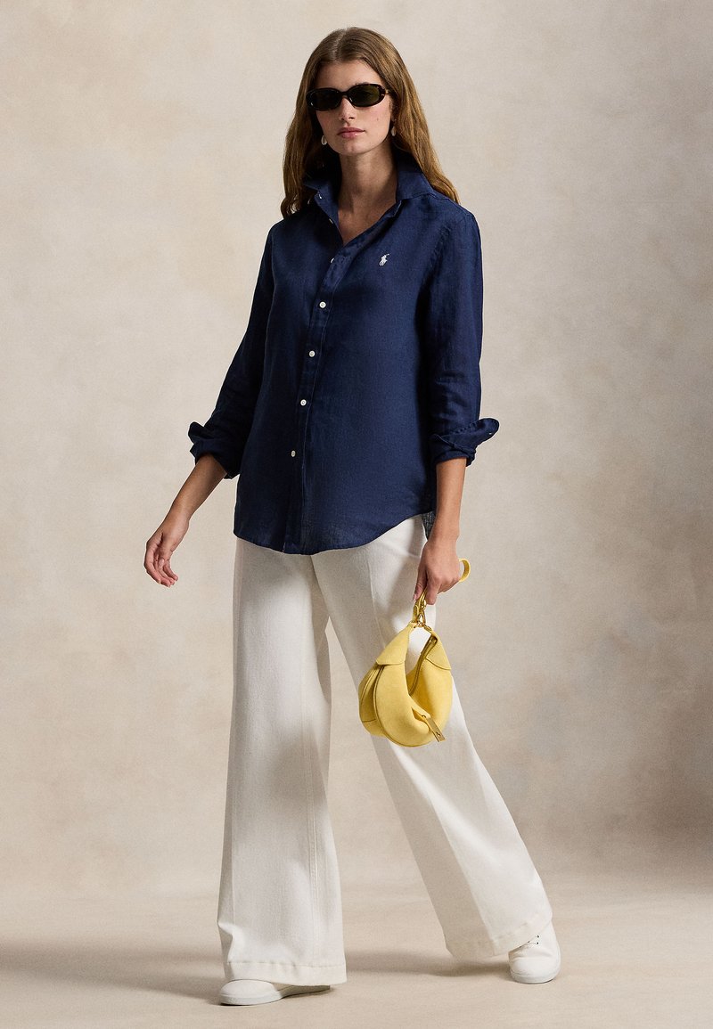 Marinblå linneskjorta med uppkavlade ärmar, vita jeans med vida ben och en gul handväska. Modellen bär svarta solglasögon och står upp.