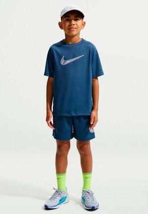 Niño de pie con camiseta y pantalones cortos azules de Nike, gorra blanca, calcetines neon verdes de Nike y zapatillas grises para correr sobre fondo liso.