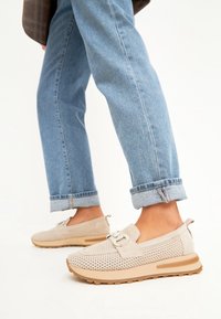 Bottines beiges sans lacets avec un dessus en mesh texturé, semelle en caoutchouc et accents beige clair ; portées avec un pantalon en denim bleu clair retroussé.