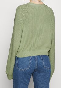 Olivgrön stickad tröja med ribbad nederkant och ärmslut, har en avslappnad passform och raglanärmar, ihop med blå högmidjade jeans.