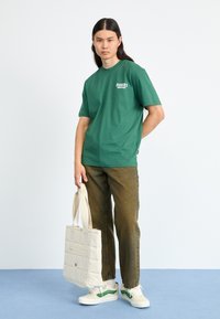 Zeleni pamučni majica s "Dickies" logotipom, uparen s smešnim izblijeđenim trapericama, bijelim tenisicama s zelenim akcentima i prešanim bež torbom.