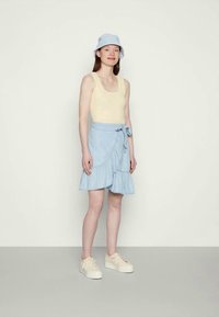 Jovem mulher de pé, vestindo uma camisola amarela, uma saia envolvente ruffled azul claro, um chapéu de balde combinando e ténis plataforma cor creme.