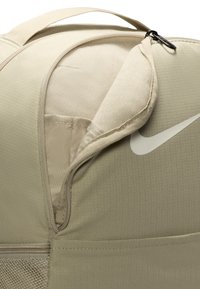 Bolsa de deporte de nailon beige con un bolsillo con cremallera, forma redondeada, textura suave y un logo blanco. Incluye un asa superior resistente para llevar.