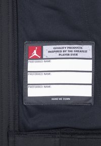 Étiquette en tissu noir avec des étiquettes blanches pour les noms ; présente du texte rouge et gris, y compris le logo "Jordan" et "HAND ME DOWN" en bas.