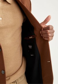 Bruine blazer met een getextureerde buitenkant en een gladde, donkere voering. Beige gebreid shirt eronder. Label aan de binnenkant met "DeFacto STUDIO," maat 50, slim fit.