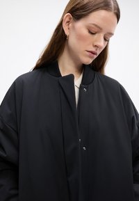 Zwarte bomberjack met een gladde, matte afwerking, voorzien van een opstaande kraag, voorste drukknoopsluiting en ruime mouwen. Minimalistisch ontwerp.