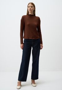 Haut long à col montant texturé marron, associé à un jeans large bleu foncé, accentué par des talons de couleur claire.