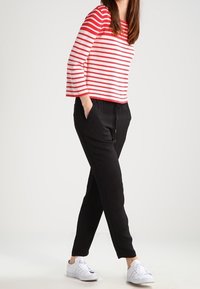 Femme portant une chemise à rayures rouges et blanches à manches longues, un pantalon noir avec cordon de serrage, des baskets blanches, les mains dans les poches, se tenant devant un fond uni.