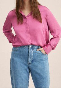 Femme portant un chemisier rose à manches longues rentré dans un jean en denim bleu clair taille haute, une main dans sa poche.
