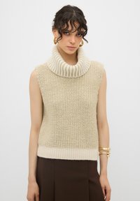 Maglione smanicato color crema in maglia con un ampio colletto a costine e corpo tessuto. Presenta un orlo a costine in vita.