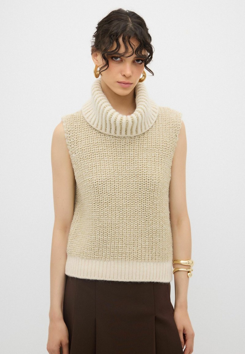 Maglione smanicato color crema in maglia con un ampio colletto a costine e corpo tessuto. Presenta un orlo a costine in vita.