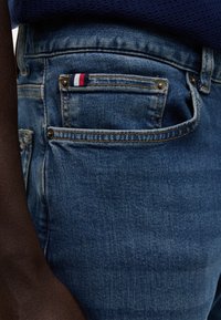 Gros plan sur la poche d’un jean en denim bleu avec une petite étiquette en tissu rouge, blanche et bleue, portée par une personne à la peau foncée et un haut bleu marine.