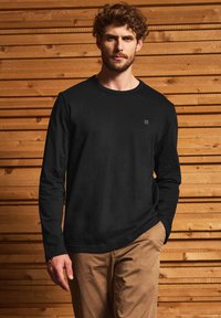 Camiseta de manga larga negra de algodón con cuello redondo, que presenta un pequeño logo en el pecho. Combinada con chinos beige frente a un fondo de madera.